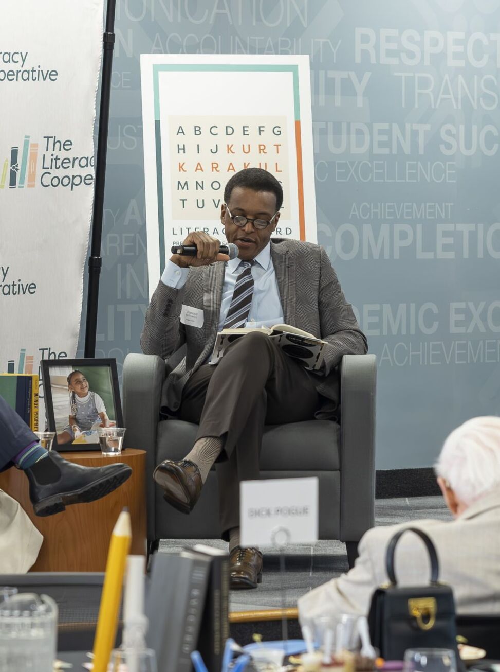 043026_230 Two men in suits sit on a panel stage, one speaking into a microphone, with The Literacy Cooperative banner in the background and an attentive audience nearby.